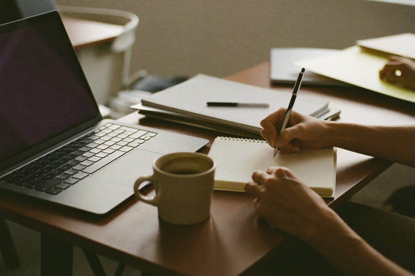 Mãos anotando em um caderno de papel ao lado de um notebook aberto sobre a mesa de um escritório.