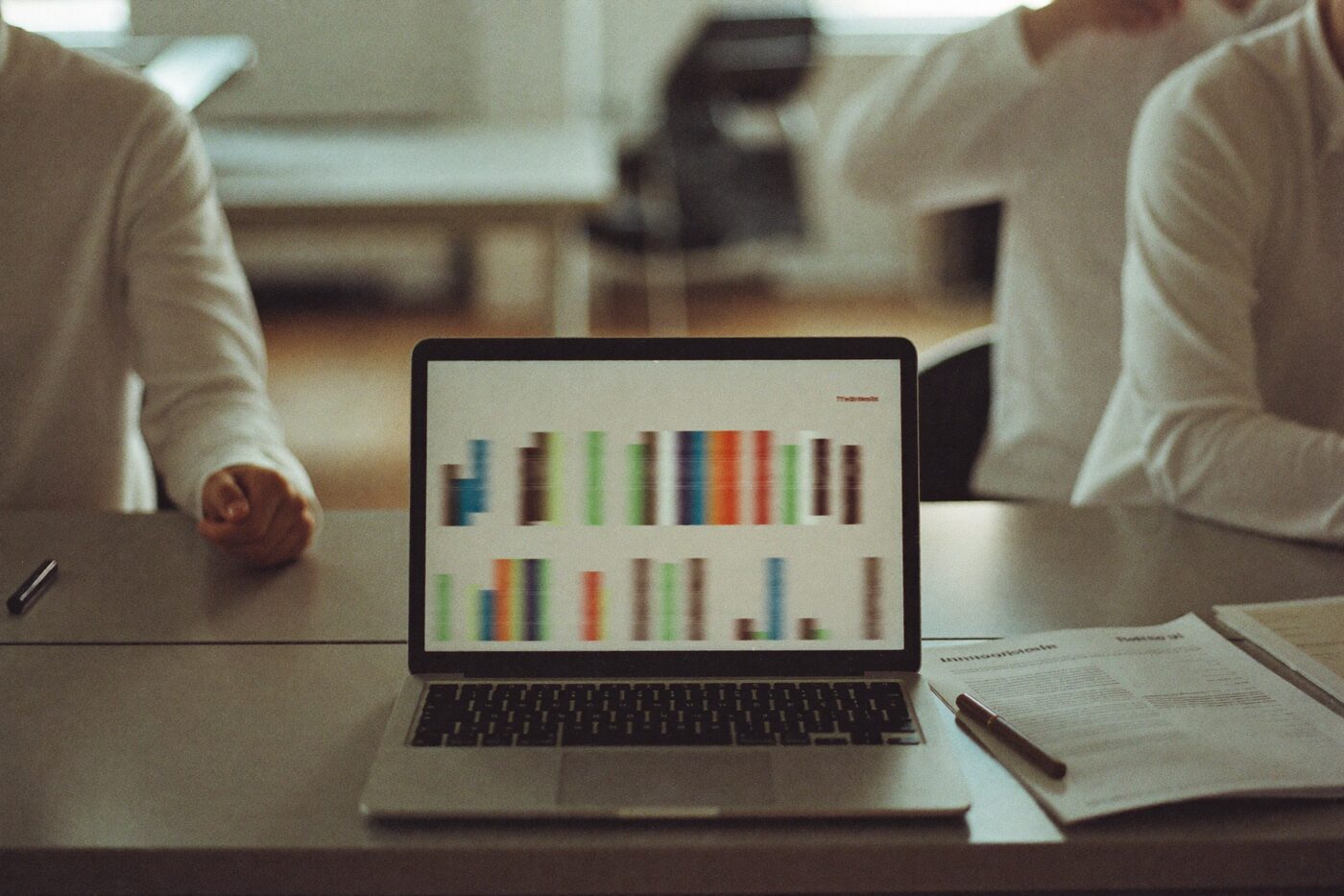 Notebook aberto sobre uma mesa exibindo gráficos, ao lado de um relatório impresso e uma caneta.