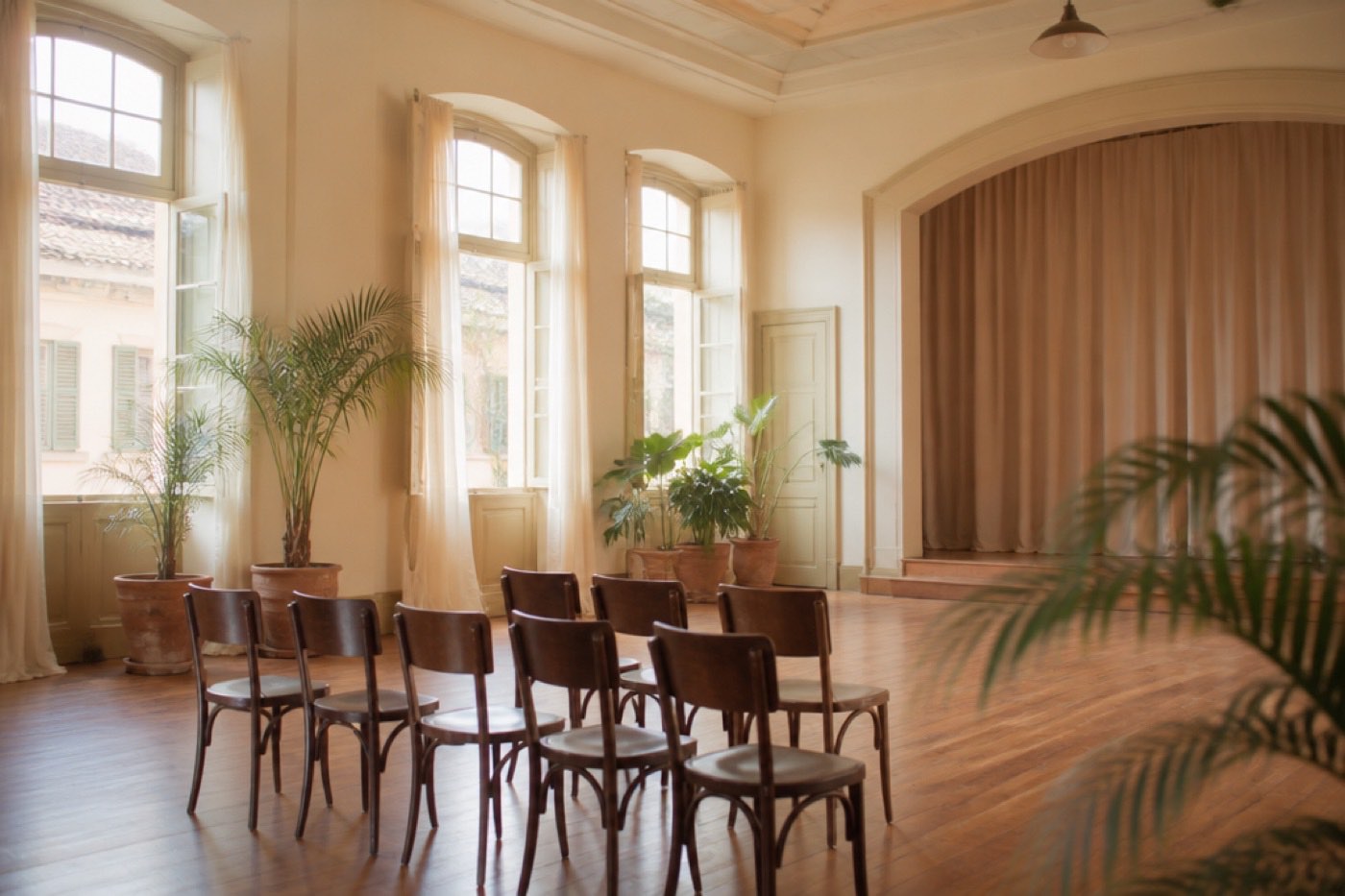 Sala de um espaço cultural com cadeiras de madeira enfileiradas diante de um pequeno palco